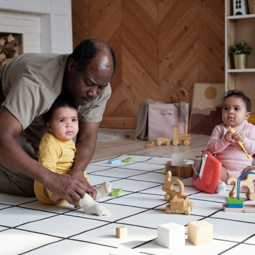Mature African American man spending time at home with his twin daughters watching them playing with toys and taking care of them
