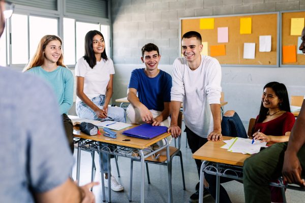 Male teacher and teenage young students talking during a break in classroom - Happy group of diverse millennial people hanging out with tutor in high school