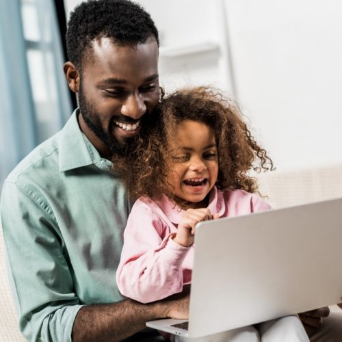 smiling african american child sitting with father on couch and using laptop
